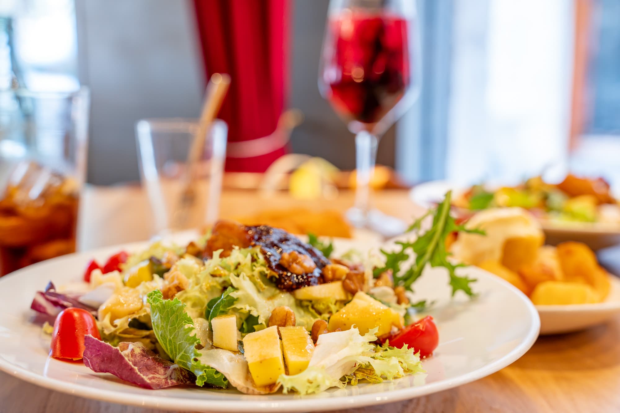 A plate of leaves with mango, cherry tomatoes and walnuts on a restaurant table, a glass of sangria and a second dish behind it, lit by daylight from a window